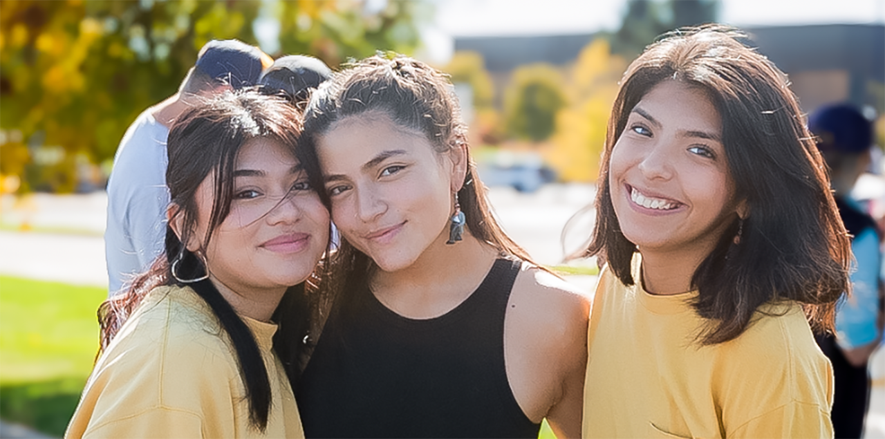 Three college students smiling 和 st和ing together outside during an event to celebrate Hispanic/Latinx communities